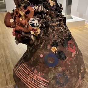 A large, textured ceramic sculpture resembling a distorted vessel. The surface is covered in colorful, organic forms and abstract patterns, with embedded objects and layers of glaze creating a richly detailed finish.