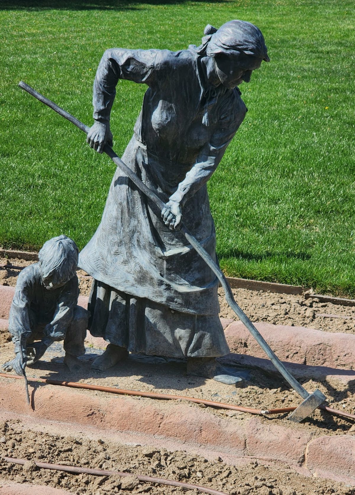 Full view of a pioneer woman hoeing in a garden with a young child beside her planting, both cast in bronze, depicting everyday frontier life.