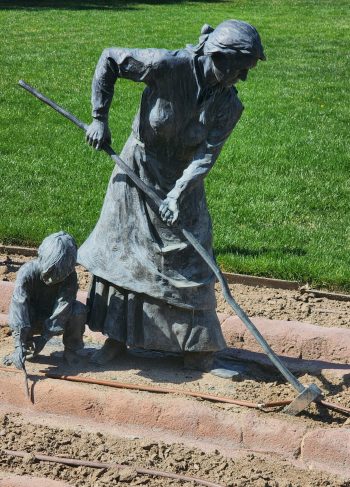 Full view of a pioneer woman hoeing in a garden with a young child beside her planting, both cast in bronze, depicting everyday frontier life.