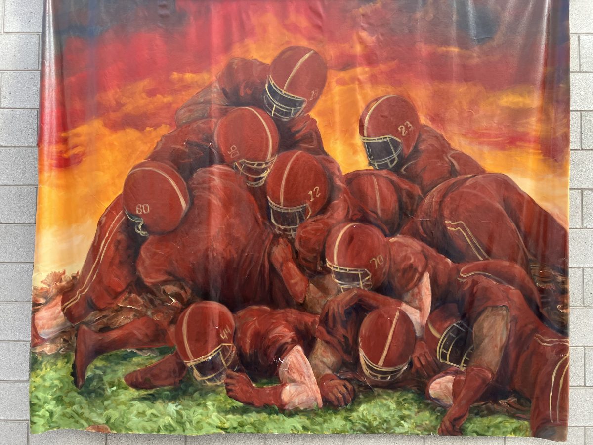 A large fabric banner depicting a pile of football players in red uniforms and helmets against a vivid orange and red sunset sky. The players are tangled together on a green field, their jersey numbers visible.