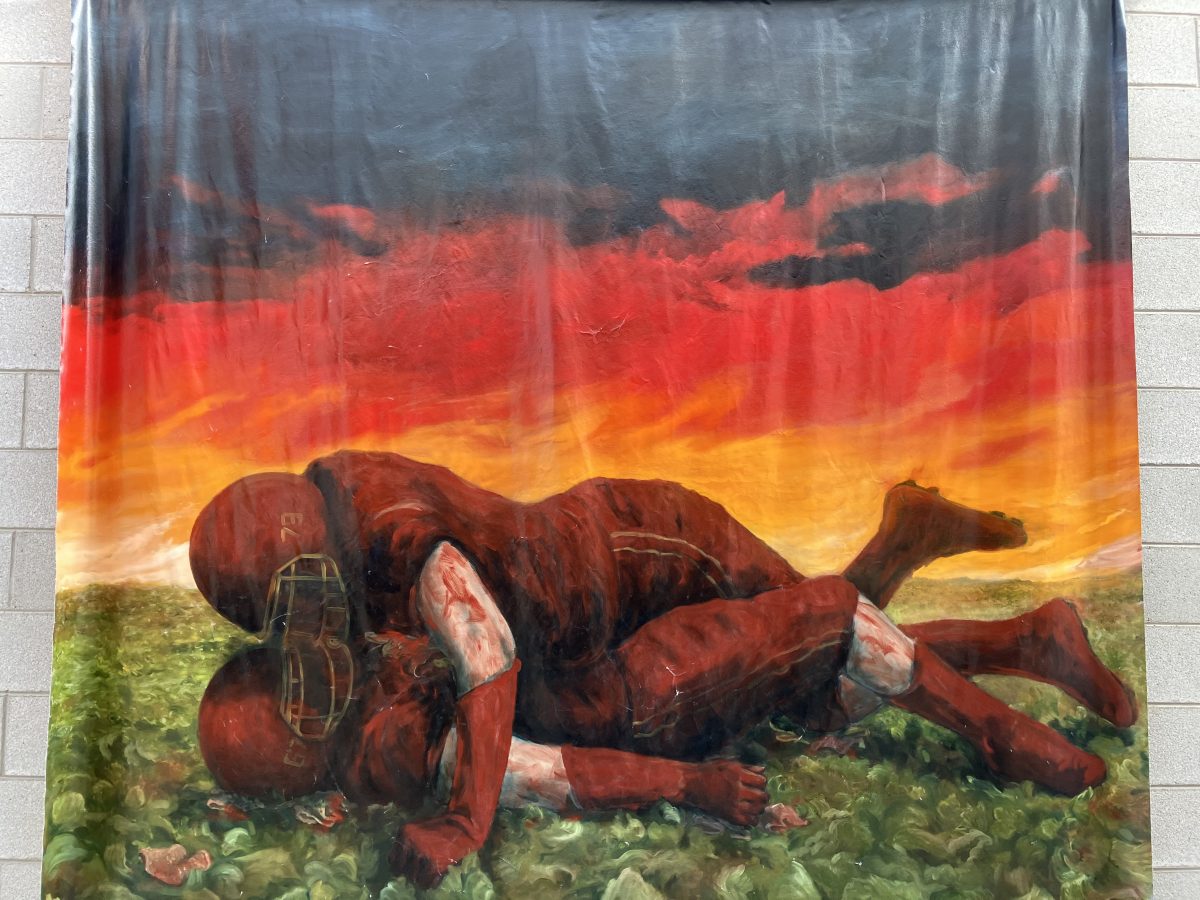 A fabric banner showing two football players in red uniforms lying on the grass under a dramatic, dark sunset sky. One player is on top of the other in an intimate, almost tender pose, creating a striking contrast with the violent sport.