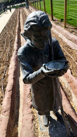 Bronze statue of a pioneer girl holding a bowl, standing in a furrowed garden bed, representing early settler life and labor.
