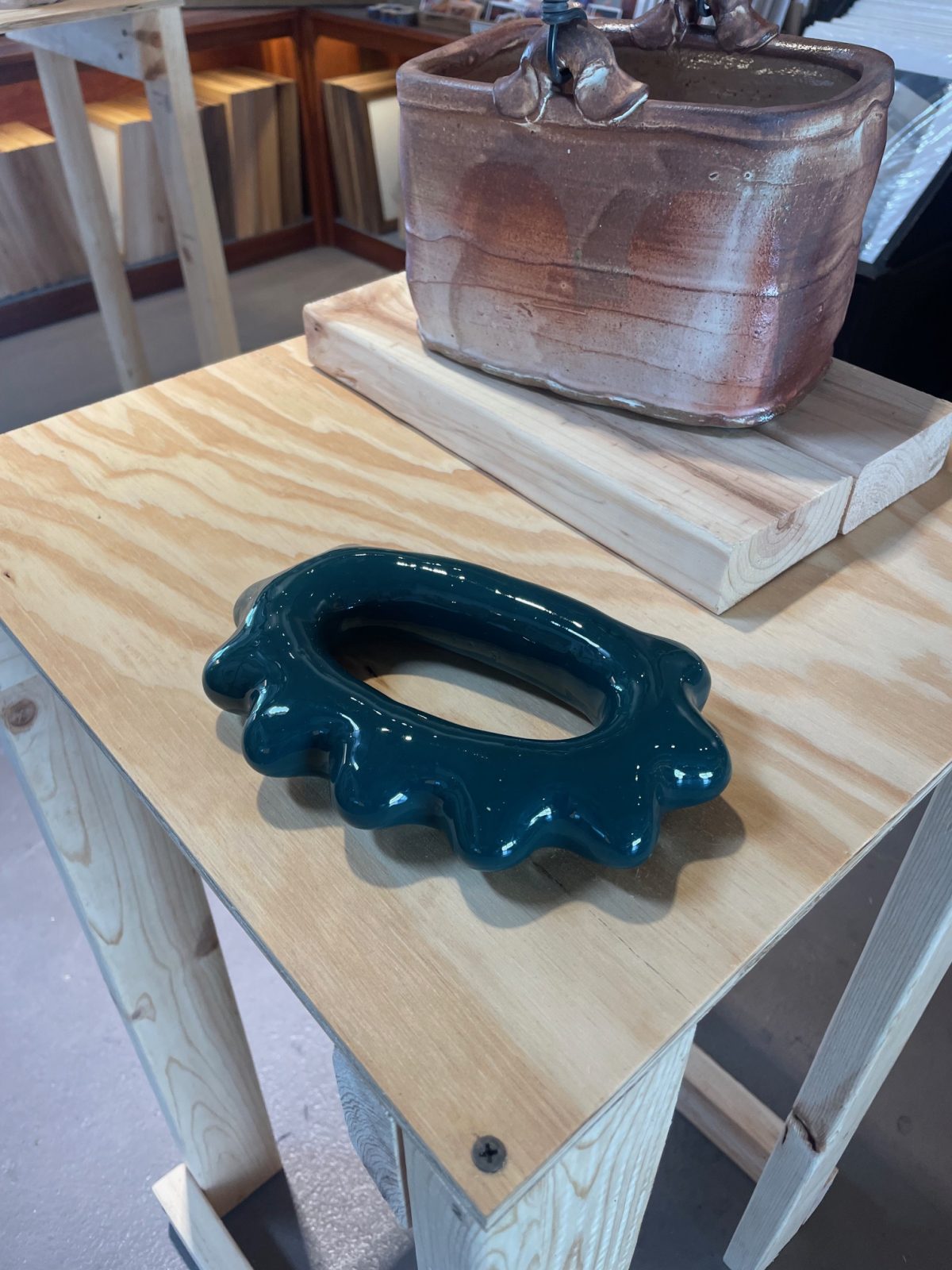 Glossy, dark green ceramic sculpture with an irregular, wavy-edged oval form, resembling a hand-held massager, displayed on a light wooden table in a gallery setting.