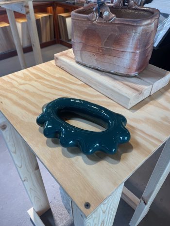 Glossy, dark green ceramic sculpture with an irregular, wavy-edged oval form, resembling a hand-held massager, displayed on a light wooden table in a gallery setting.