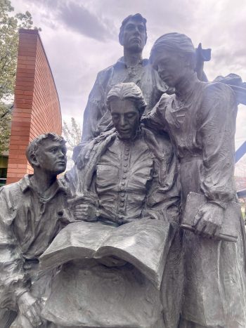 A bronze sculpture of five figures arranged closely together. An older woman at the center holds an open book, surrounded by a young boy, a woman with a second book, and two men, all engaged in the act of reading or reflection.