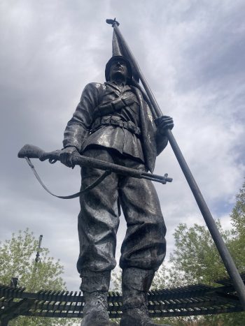 A bronze statue of a soldier holding a rifle and a tall flagpole, viewed from below. The figure stands at attention, wearing a helmet and uniform, with trees and a cloudy sky in the background.