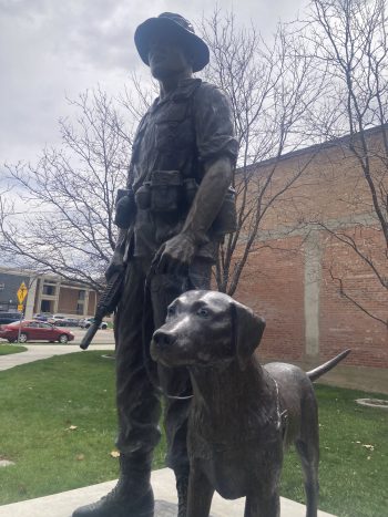A bronze statue in Tooele depicting a soldier in uniform standing beside a loyal service dog. The soldier wears a helmet and holds a rifle, while the dog stands alert at his side. Leafless trees and a red brick building are visible in the background.