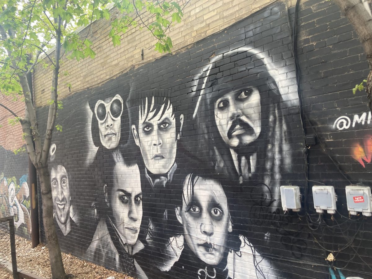 A black-and-white mural depicting several of Johnny Depp’s most iconic film characters, including Captain Jack Sparrow, Edward Scissorhands, and Willy Wonka, painted on a brick wall under leafy trees.