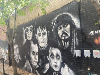 A black-and-white mural depicting several of Johnny Depp’s most iconic film characters, including Captain Jack Sparrow, Edward Scissorhands, and Willy Wonka, painted on a brick wall under leafy trees.