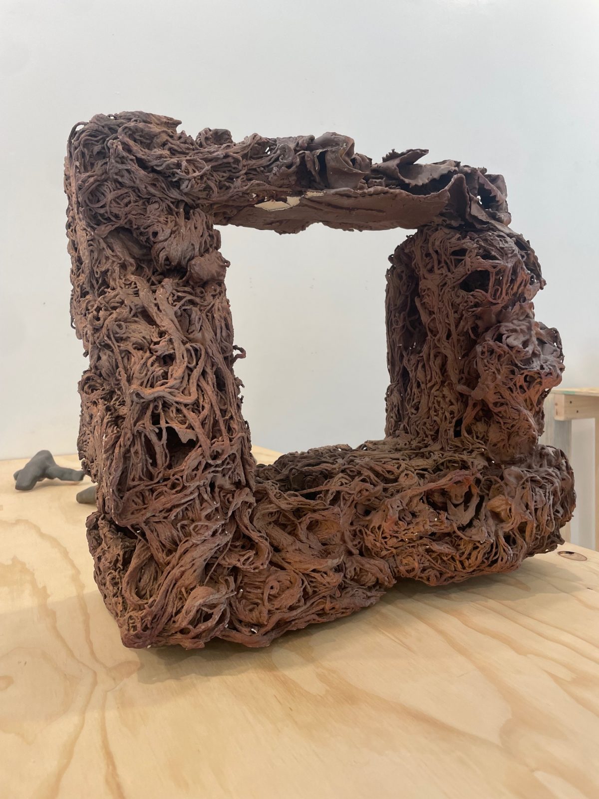 extural ceramic sculpture resembling a hollow, organic frame with a heavily layered and tangled surface, made from rich reddish-brown clay, displayed on a wooden tabletop.