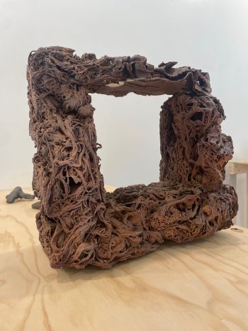 extural ceramic sculpture resembling a hollow, organic frame with a heavily layered and tangled surface, made from rich reddish-brown clay, displayed on a wooden tabletop.