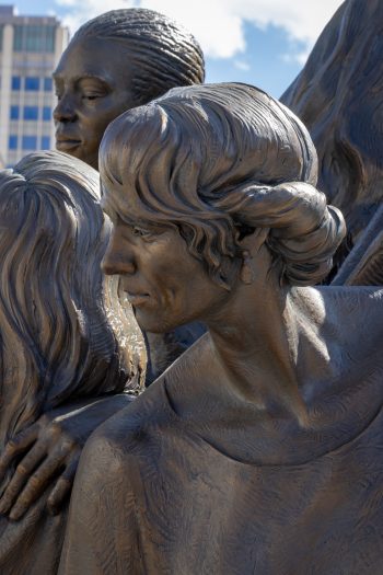A close-up of The Five Wise Virgins sculpture highlights the detailed facial features and textured hair of two women. One looks gently to the side with her hair styled in a braid, while the other, in the foreground, has short, wavy hair and wears a single teardrop earring, adding individuality to each figure.
