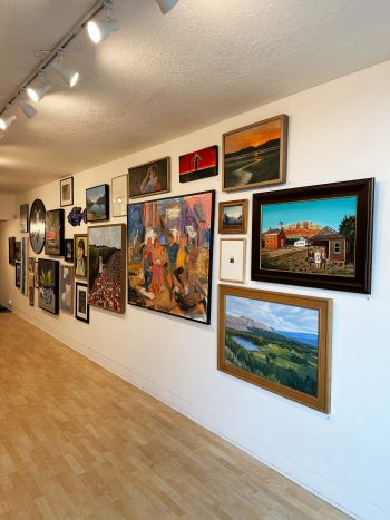 A gallery wall displaying an eclectic mix of framed paintings and drawings, including landscapes, portraits, abstract works, and small mixed-media pieces, all arranged salon-style on a white wall with a wood floor.”