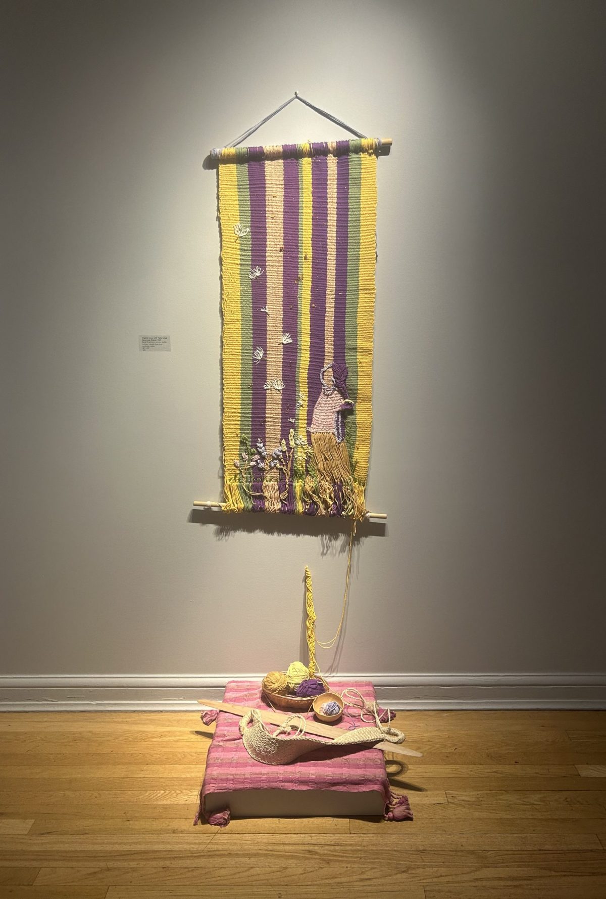 A handwoven textile with bright vertical stripes in purple, yellow, green, and peach. At the bottom, a figure kneels beside a blooming field. The work is mounted with weaving tools and yarns placed on a pink cushion beneath it, suggesting an in-process loom.