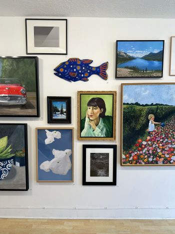 A section of gallery wall with an eclectic mix of artwork including a bright blue sculptural fish, a woman’s portrait, abstract paper-like forms, a vibrant flower garden scene with a child, and various landscape paintings.