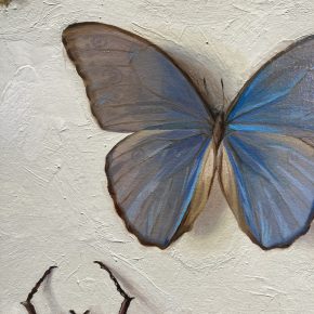 Close-up of a blue butterfly painted by Nicole Parish, showing intricate detail in the wings and lifelike coloration against a textured off-white background.