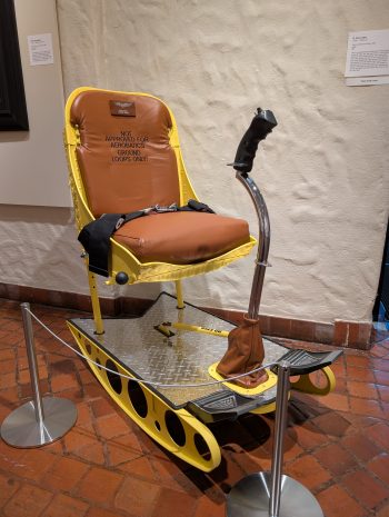 A bright yellow aviation-themed seat with brown cushion labeled “Not approved for aerobatics, ground loops only!”, complete with safety harness, joystick, and metal base.