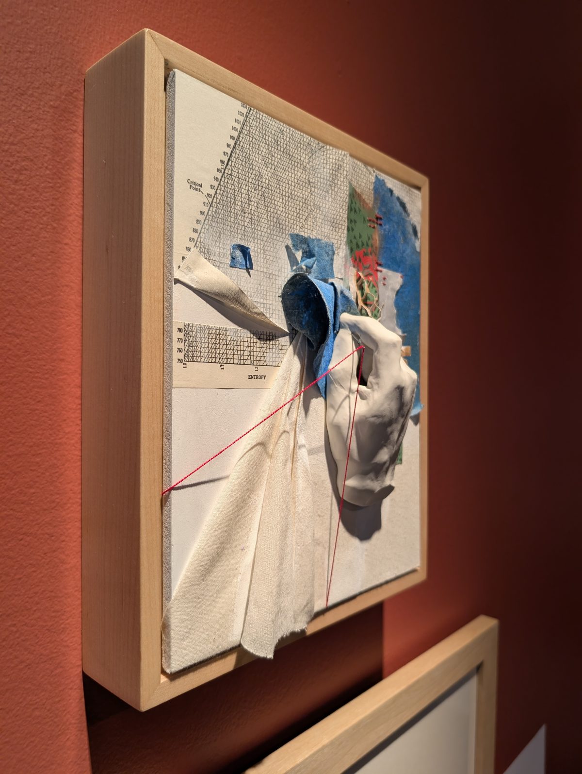 A framed artwork combining fabric, graph paper, blue and red shapes, and a three-dimensional white hand, with a grid and entropy chart in the background.