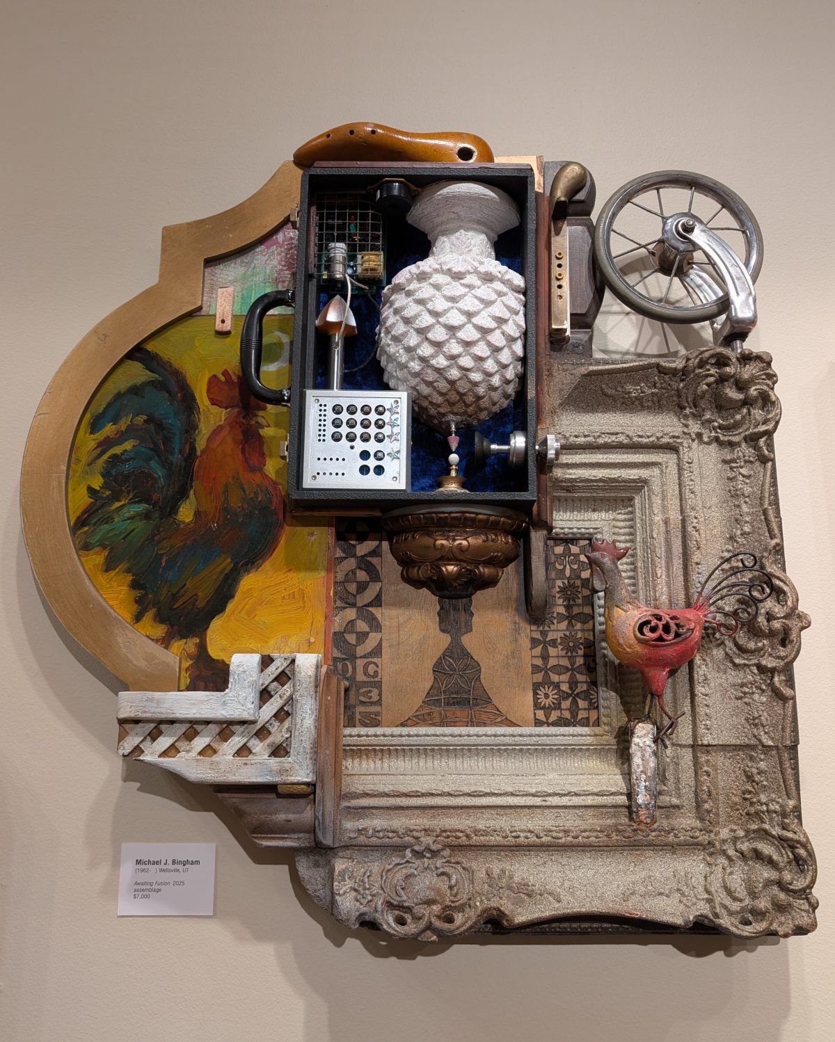 A framed sculptural assemblage combining a rooster painting, carved wooden shapes, a vase, mechanical components, and a small red rooster figurine, all arranged on an ornate backing.