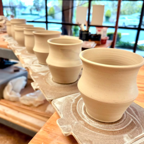 A row of freshly thrown clay mugs lined up on bats atop a work table in a pottery studio, with natural light streaming through large windows.