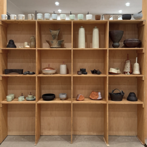A wooden shelving unit displaying a diverse collection of ceramic vessels and sculptures, including mugs, teapots, and decorative vases in various styles and finishes.