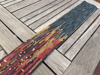A long knitted scarf with color shifts from dark to bright, interspersed with small, multicolored gemstones, laid out on a wooden table.