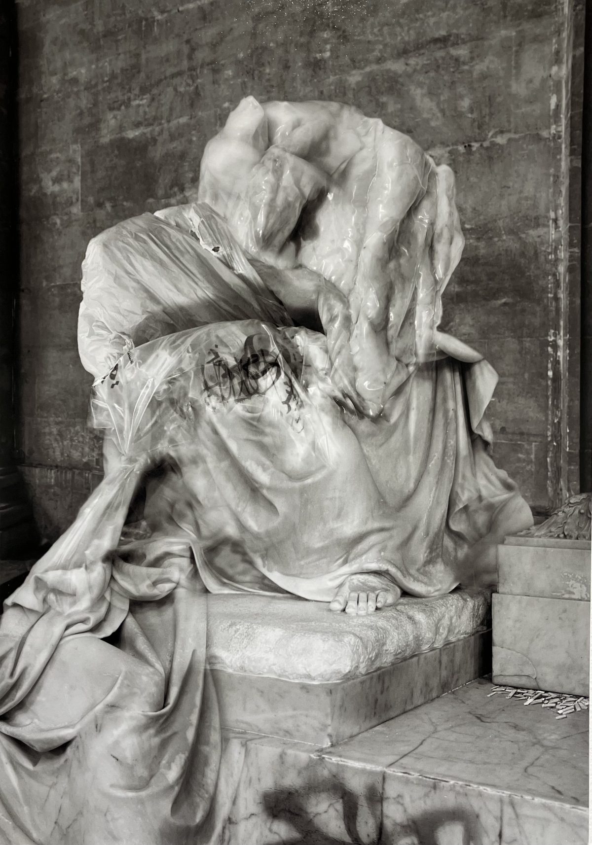 Photograph of a veiled classical statue seated on a marble base, partially obscured by translucent plastic wrapping and graffiti marks.