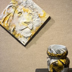 A wall-mounted relief of a lion in yellow-and-white glass hangs above a similarly colored blown glass vessel with swirling patterns.