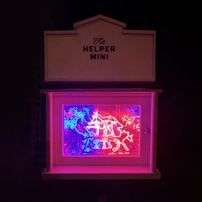 White miniature gallery box with “The Helper Mini” sign, glowing at night with layered neon lights in red, pink, and blue, forming stylized figures and shapes.