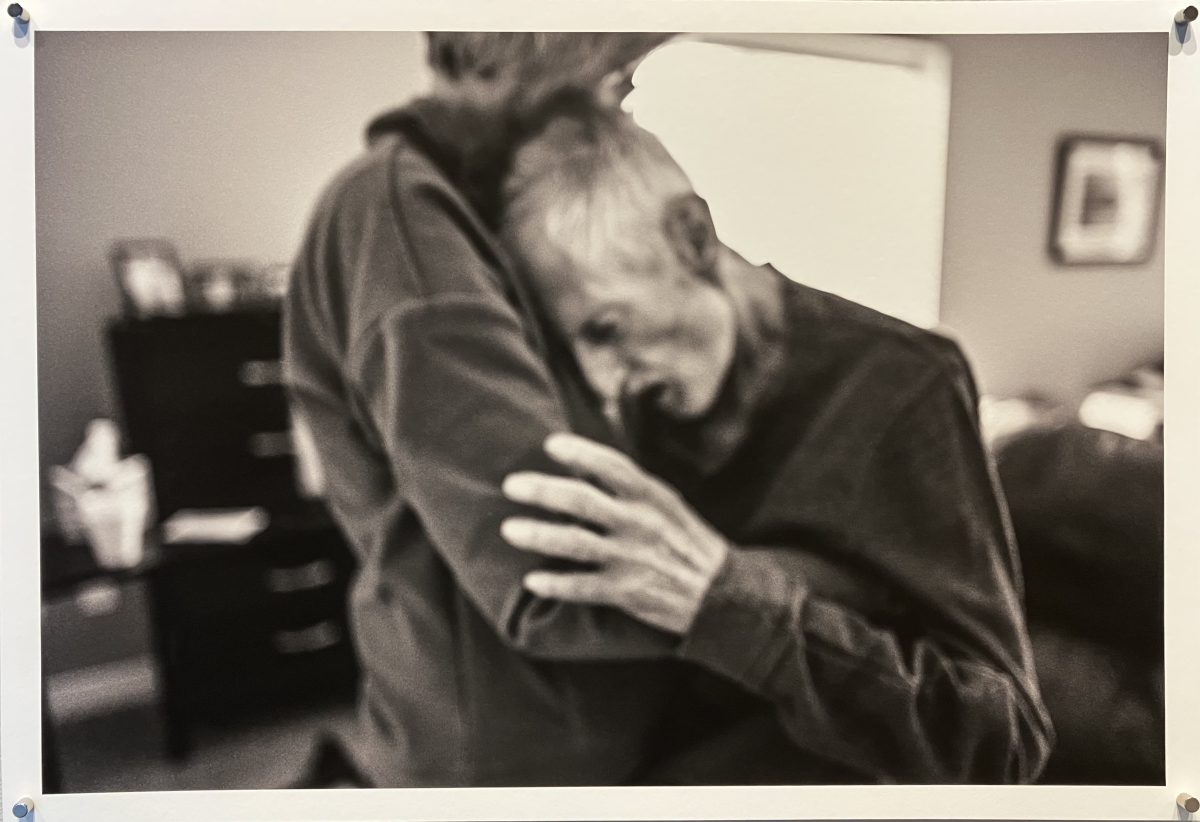 An elderly man buries his head into a woman’s shoulder as she embraces him, their bodies conveying grief and intimacy in a blurred, emotional image.