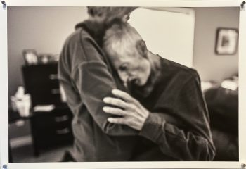 An elderly man buries his head into a woman’s shoulder as she embraces him, their bodies conveying grief and intimacy in a blurred, emotional image.