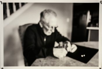 A blurred black-and-white image of an elderly man sitting at a kitchen table, his head bowed as he looks down at a few pills in front of him.