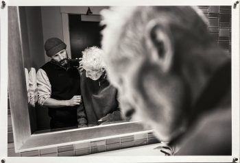 A man photographs his elderly father in the bathroom mirror, steadying him as the father looks down, while the blurred profile of the father dominates the foreground.