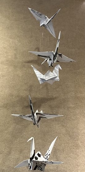 A vertical mobile of origami paper cranes folded from sheets covered in black-and-white calligraphy, suspended in flight against a neutral background.