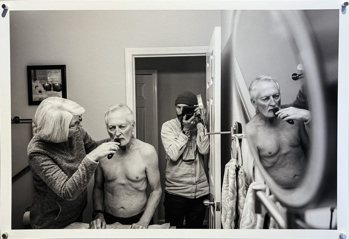 A woman helps an elderly shirtless man shave in the bathroom while their son photographs the moment in the mirror, capturing layers of reflection.