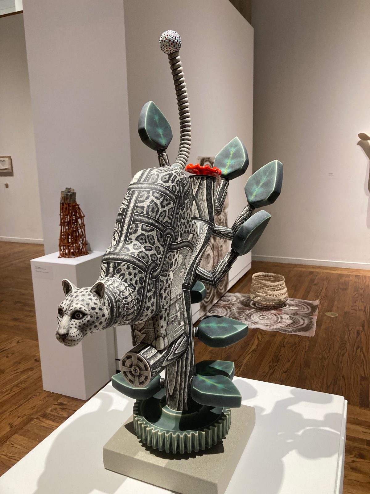 A ceramic sculpture of a jaguar with mechanical tubing, cactus-like leaves, and a dotted antenna emerging from its back, displayed in a gallery setting.