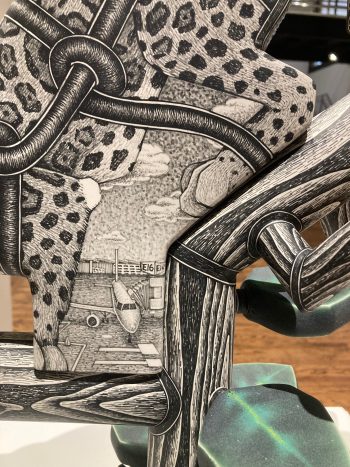 Close-up view of a ceramic sculpture’s surface showing leopard spots, pipes, and a finely drawn scene of an airplane parked at an airport gate.