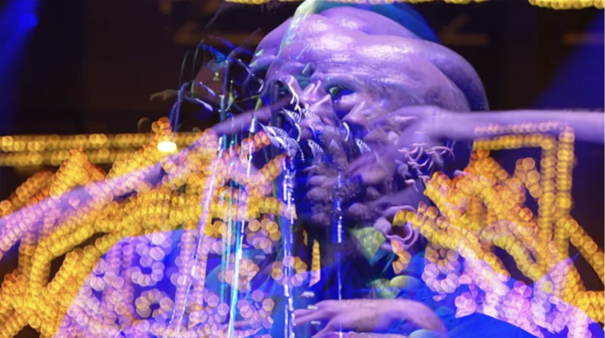 Color-saturated photographic image of a performer with blurred hands and face against glowing yellow and blue lights, creating an energetic, kaleidoscopic effect.
